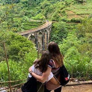 Two women from a WeRoad group trip embrace while looking at a stone arch bridge in a lush, green valley.