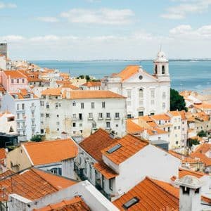 Una vista dall'alto di una città con tetti in tegole arancioni e edifici bianchi che si affacciano su un grande specchio d'acqua sotto un cielo azzurro.