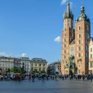 Una folla di persone passeggia in una storica piazza cittadina di fronte a una basilica di mattoni con due torri, in una giornata di sole.