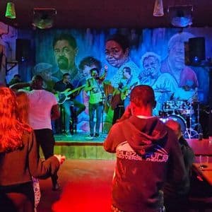 In einem schummrigen Club spielt eine Live-Band auf einer Bühne mit einem großen Wandgemälde im Hintergrund vor Publikum.
