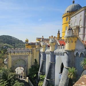 Un château jaune et bleu coloré, doté de tours et de remparts, avec des visiteurs sur ses chemins de ronde, sur fond de colline verdoyante et boisée.