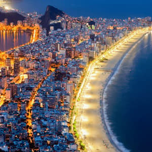 A nighttime aerial view of a brightly lit coastal city with a long, illuminated beach curving along the dark ocean.