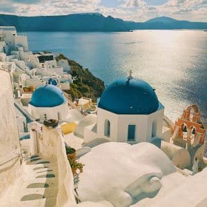 Chiese con cupole azzurre ed edifici bianchi sorgono su una scogliera che si affaccia su un mare scintillante, con montagne lontane sotto un cielo azzurro.