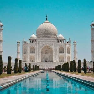 Veduta frontale del mausoleo Taj Mahal in marmo bianco, riflesso in una lunga e stretta vasca sotto un cielo azzurro brillante.