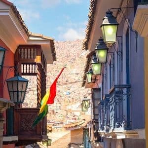 Uno scorcio di una stretta via coloniale con muri colorati, lanterne e una bandiera boliviana, che si affaccia su una città costruita su una collina.