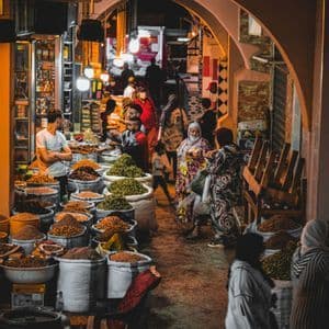 La gente pasea por un callejón de mercado cálidamente iluminado por la noche, pasando puestos con grandes sacos de especias y hierbas de colores.