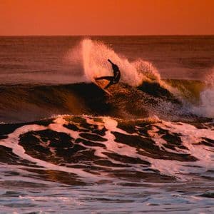 Un surfista in silhouette cavalca una grande onda oceanica, sollevando spruzzi contro un cielo arancione brillante al tramonto.