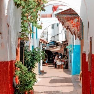 Una vista de un callejón estrecho y soleado con arcos blancos, paredes rojas y azules vibrantes, y puestos de mercado a lo lejos.