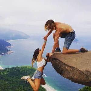 Un uomo è inginocchiato sul bordo di una scogliera rocciosa, tenendo la mano di una donna appesa sotto, con una suggestiva costa sullo sfondo.