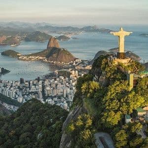 La statua del Cristo Redentore si erge su una montagna boscosa che domina una città costiera, una baia e altre montagne al tramonto.
