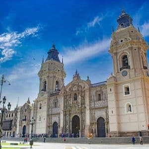 Una cattedrale ornata con due grandi campanili si erge in una piazza cittadina sotto un cielo azzurro e parzialmente nuvoloso.