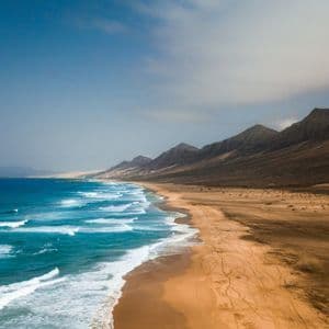 Una vista aerea di onde turchesi che si infrangono su una vasta spiaggia sabbiosa ai piedi di una catena montuosa.