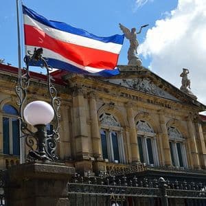 Le drapeau du Costa Rica flotte sur un mât devant la façade en pierre ornée du bâtiment du Théâtre National, sous un ciel bleu nuageux.