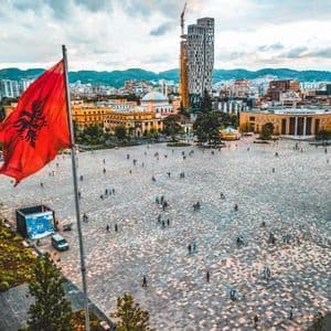 An Albanian flag waves over a large, bustling city square filled with people, with a city skyline in the background.