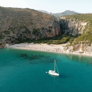 An aerial view of a white sailboat anchored in a turquoise sea bay next to a secluded beach surrounded by steep cliffs.