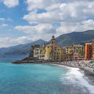 Un colorato villaggio costiero con una chiesa su una scogliera, una spiaggia di ciottoli affollata e acqua turchese.