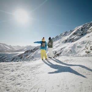 Dos personas de un viaje de grupo de WeRoad están de pie en la cima de una montaña nevada, señalando el paisaje bajo un sol brillante