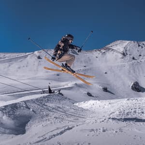 Un esquiador salta en el aire desde una rampa de nieve en la ladera de una montaña bajo un cielo azul despejado