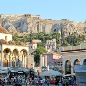 Ruinas antiguas se alzan sobre una gran colina rocosa, dominando una bulliciosa plaza de la ciudad con edificios históricos abovedados y multitudes de gente.