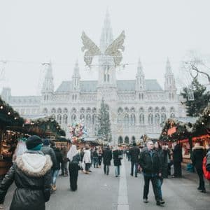 Una folla di persone passeggia in un mercatino di Natale all'aperto, con bancarelle decorate, di fronte a un grande edificio storico.