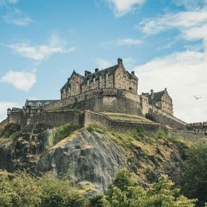 Ein großes Steinschloss thront auf einem felsigen, grünen Hügel, von unten sichtbar unter einem teils bewölkten blauen Himmel.