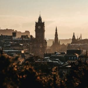 A historic cityscape with a prominent clock tower and spires silhouetted against a hazy, warm sunrise, viewed from behind dark treetops.