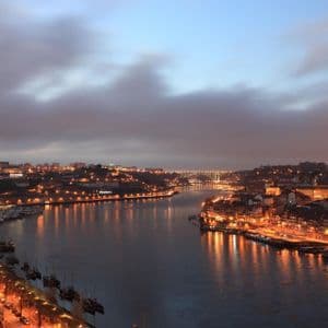Paisaje urbano iluminado a lo largo de un río ancho al anochecer, con las luces de la ciudad reflejándose en el agua y un puente al fondo.