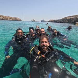 Un gruppo WeRoad in gita subacquea sorride per un selfie mentre galleggia in acque limpide e turchesi vicino a una costa rocciosa.
