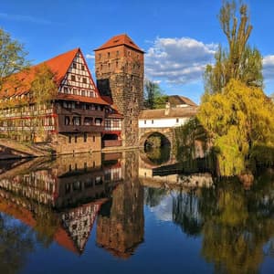 Edifici medievali a graticcio e una torre di pietra perfettamente riflessi in un fiume calmo sotto un cielo azzurro.