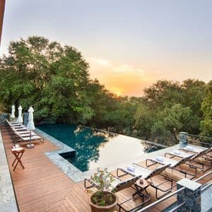 Ein Infinity-Pool mit Liegestühlen auf einer Holzterrasse, mit Blick auf einen Wald bei Sonnenuntergang.