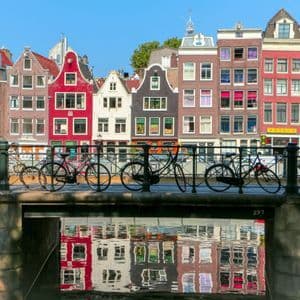Biciclette parcheggiate su un ponte che attraversa un canale, con case tradizionali colorate e i loro riflessi nell'acqua sullo sfondo.