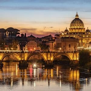 Una gran basílica iluminada y un puente de piedra arqueado se reflejan en un río durante un atardecer colorido.