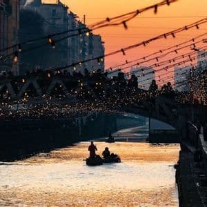 A small boat floats on a city canal as people walk across a bridge adorned with string lights against a sunset sky.