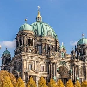 Une cathédrale ornée avec de grands dômes verts et une haute tour de télévision en arrière-plan, vue au-dessus d'arbres d'automne jaunes.