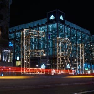 Grandi lettere illuminate che formano 'BERLINO' su una strada cittadina di notte, con scie luminose dal traffico e un edificio moderno sullo sfondo.