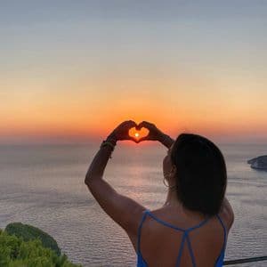 Eine Frau von hinten gesehen formt mit ihren Händen ein Herz, das die untergehende Sonne über dem Meer umrahmt.