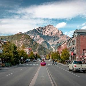 Una strada ampia in un paese di montagna con auto e pedoni, che conduce verso un'imponente vetta rocciosa sotto un cielo parzialmente nuvoloso.