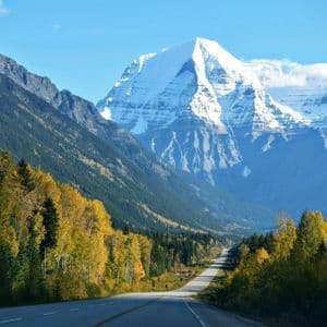 Una strada asfaltata attraversa una valle fiancheggiata da alberi autunnali verso un'imponente montagna innevata sotto un cielo azzurro.