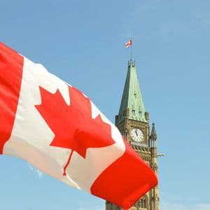 La bandiera canadese sventola su un pennone davanti a una torre dell'orologio in pietra contro un cielo azzurro chiaro.
