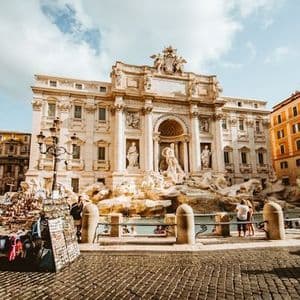 Una gran fuente ornamentada en una plaza empedrada, con turistas reunidos alrededor y un puesto de souvenirs en primer plano.