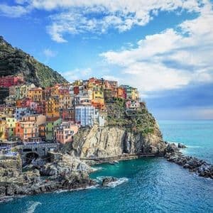 A village of colorful houses built into a rocky cliffside overlooking a turquoise sea under a blue sky with white clouds.