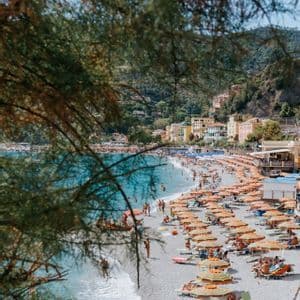 A crowded pebble beach viewed from above, filled with people relaxing under orange umbrellas next to turquoise water.