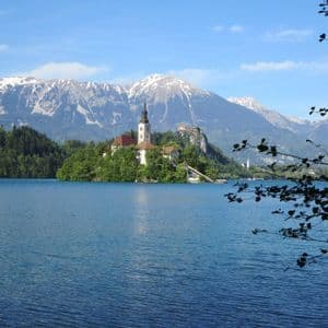 Una chiesa su una piccola isola alberata in un lago, con montagne innevate sullo sfondo sotto un cielo blu.