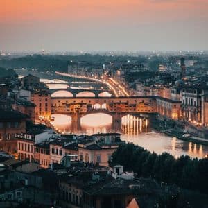 An illuminated stone bridge crosses a river in a dense cityscape at dusk, its lights reflecting on the water.