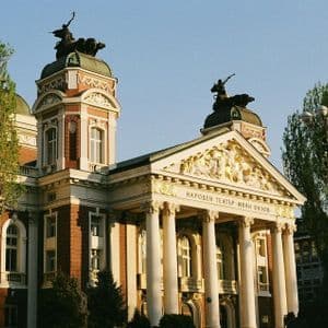 La fachada iluminada por el sol de un edificio ornamental de estilo neoclásico con grandes columnas blancas, un frontón dorado y estatuas en sus torres.