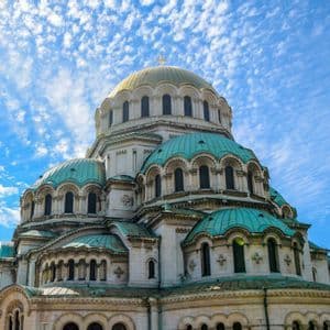 Una gran catedral con una cúpula central dorada y cúpulas turquesas más pequeñas contra un cielo azul brillante con nubes ligeras.