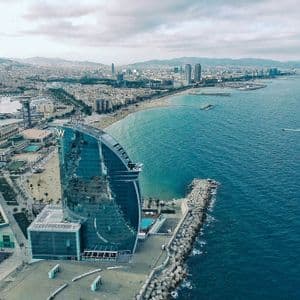 Vista aerea di una città costiera con un hotel di vetro a forma di vela sul lungomare, accanto a una lunga spiaggia sabbiosa.