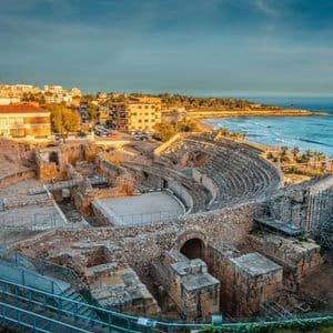 Un antico anfiteatro in pietra in rovina affacciato sul mare e una città costiera al tramonto.