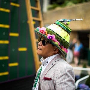 Un uomo in blazer a righe e con un elaborato cappello a tema tennis guarda un grande tabellone segnapunti verde.