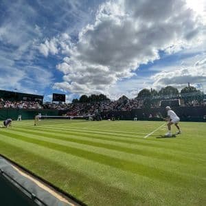 Inquadratura dal basso di una partita di tennis su campo in erba a strisce, con un giocatore in bianco pronto a giocare sotto un cielo azzurro nuvoloso.